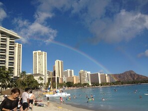 Ein Strand mit Stadtansicht im Hintergrund, einem Regenbogen und einem Boot auf dem Wasser.