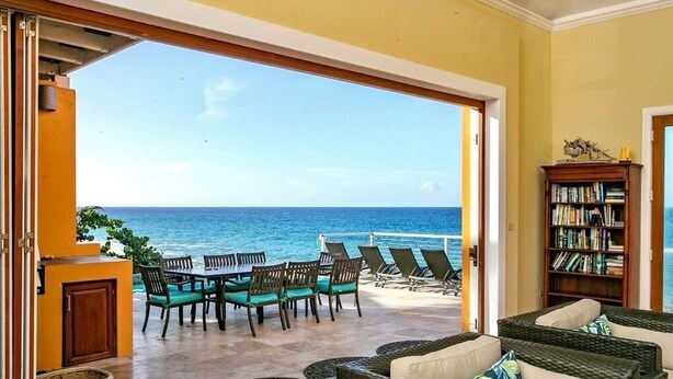 A room with a view of the ocean, a dining area with chairs and a table, and a bookshelf. - Private Gated Ocean Front Villa Pool&Tide Pool Cane Bay Beach Wifi Wedding Venue (Christiansted)