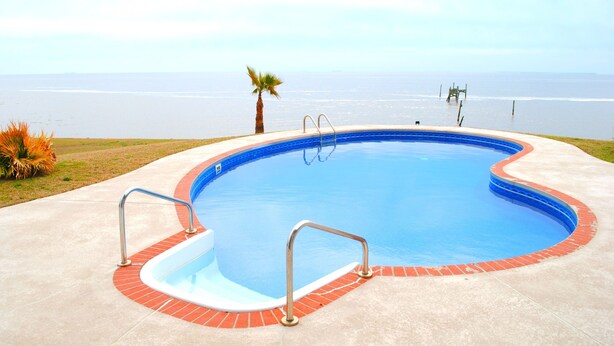 A swimming pool with a view of the ocean, surrounded by a concrete deck and a palm tree. - Gulf Front With Great Views And Pool! (Ocean Springs)