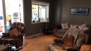 Un salon avec un fauteuil marron, un canapé et une table basse en verre. Il y a une fenêtre avec des stores, une lampe sur une table d’appoint et un tableau encadré au mur.