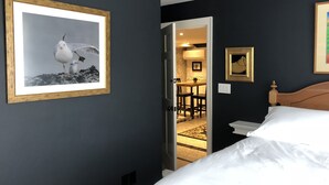 A hotel room with a bed, a framed seagull picture, a small table with chairs, and a doorway leading to a dining area.