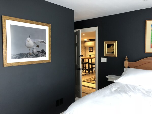 A hotel room with a bed, a framed seagull picture, a small table with chairs, and a doorway leading to a dining area.