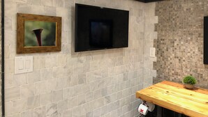 A bathroom with a tiled wall, a mounted TV, a framed picture, a wooden vanity, and a potted plant.
