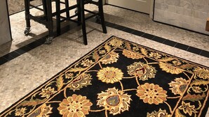 A dining area with a wooden table and chairs, a potted plant, and a patterned rug.