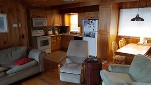 A cozy cabin-style kitchen with wooden cabinets, a microwave, stove, and a refrigerator. There is a dining area with a table and chairs. - Great Beach House - Kill Devil Hills, One Row from Oceanfront (Kill Devil Hills)