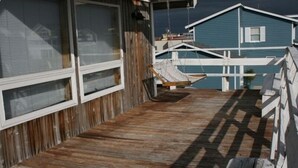 A wooden deck with a swing chair, a white railing, and a view of a blue building in the background. - Great Beach House - Kill Devil Hills, One Row from Oceanfront (Kill Devil Hills)