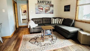 A living room with a dark sofa, a patterned rug, wooden flooring, and a ceiling with exposed beams and hanging light fixtures. - Relaxing Lakefront Cabin, Family + Lake + Dock + Sunset + Be Here, All Year Fun! (Watertown)