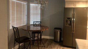 A compact kitchen with a dining table, chairs, a chandelier, a refrigerator, and a trash can.
