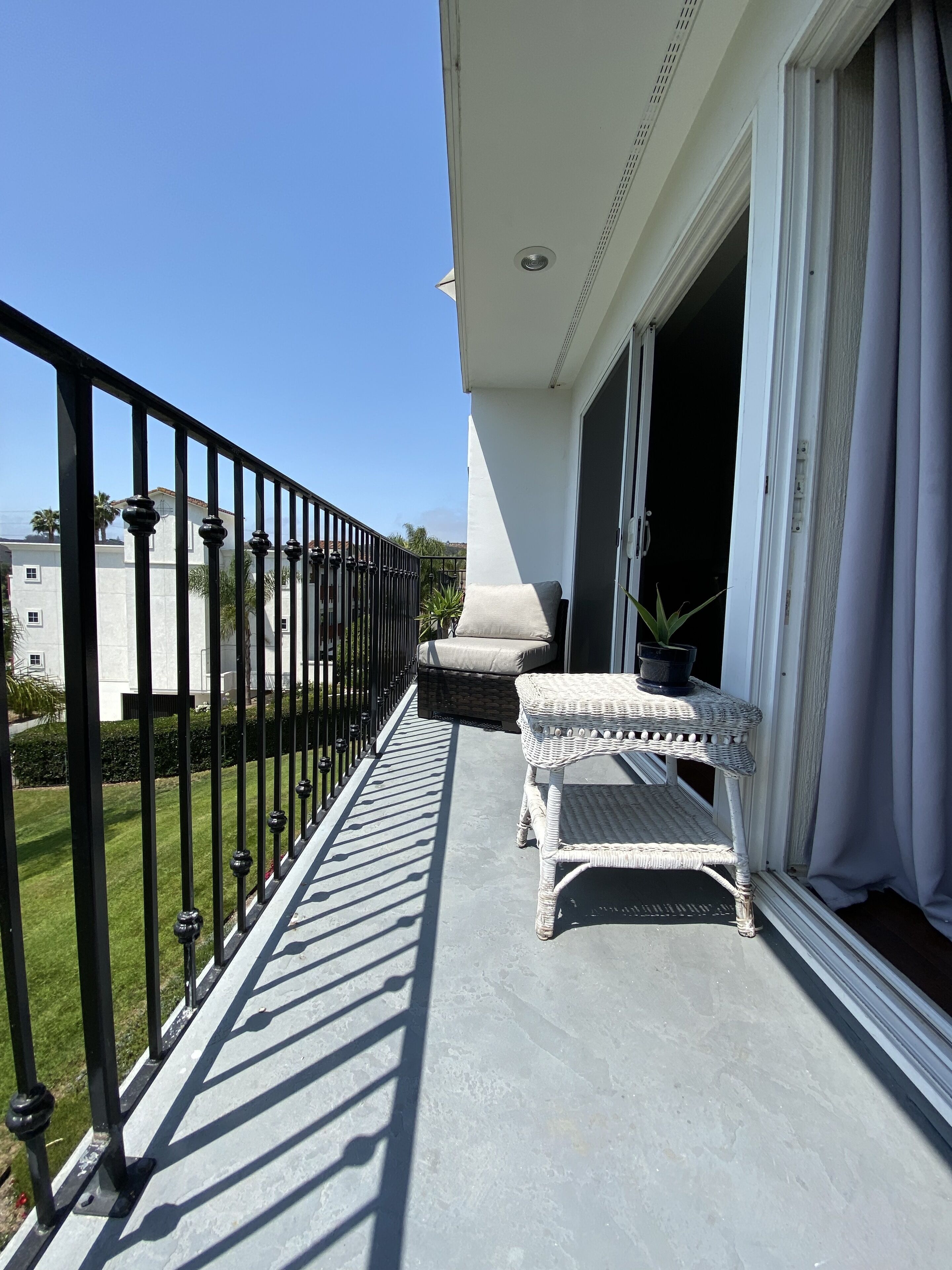 A balcony with a white wicker table and bench, a black metal railing, and a view of the surrounding area.