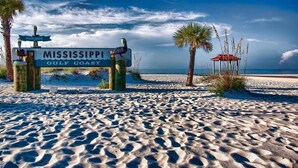A beach scene with a 'Mississippi Gulf Coast' sign, palm trees, and a beach umbrella. - Agape Bay - Sienna on the Coast Unit 102 (Gulfport)