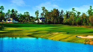 A golf course with a water body, trees, and houses in the background. - Agape Bay - Sienna on the Coast Unit 102 (Gulfport)