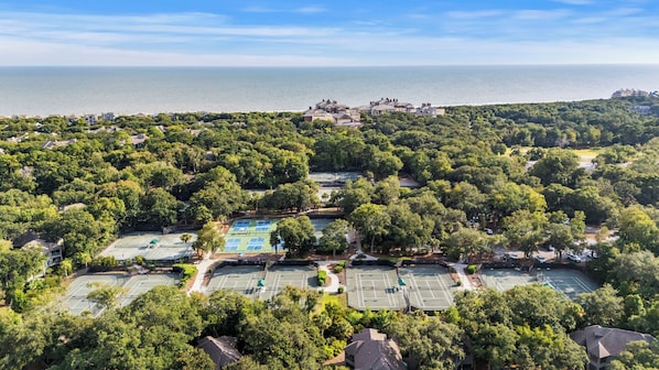 Luchtbeeld van een woongebied met meerdere tennisbanen, omgeven door weelderige begroeiing, een helder uitzicht op de zee en een verafgelegen groep gebouwen.