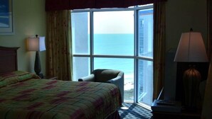 Ein Hotelzimmer mit Bett, Sessel, Schreibtisch mit Lampe und einem großen Fenster mit Meerblick.