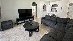 Een woonkamer met een grote flatscreentelevisie, een hoekbank, een salontafel en een plafondventilator.