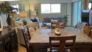 A spacious living room with a large dining table, comfortable seating, a television, and a kitchen area.