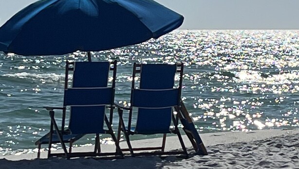 Two beach chairs under a blue umbrella on a sandy beach with a view of the ocean.