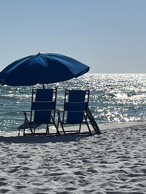 Zwei Liegestühle unter einem blauen Sonnenschirm an einem sandigen Strand mit Blick aufs Meer.