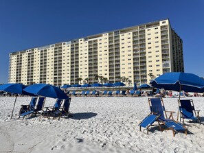 A large hotel with multiple floors, a sandy beach in front, and blue umbrellas.