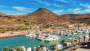 Un puerto deportivo con barcos amarrados, una carretera con vehículos y un paisaje montañoso al fondo.