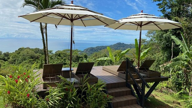 Une terrasse dotée de deux parasols, de chaises en osier et offrant une vue sur l’océan.