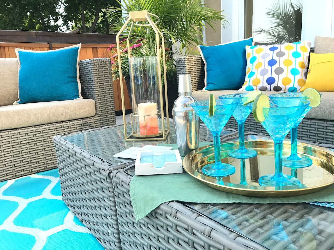 A wicker sofa with blue cushions, a glass table with drinks, a candle, and a lantern.