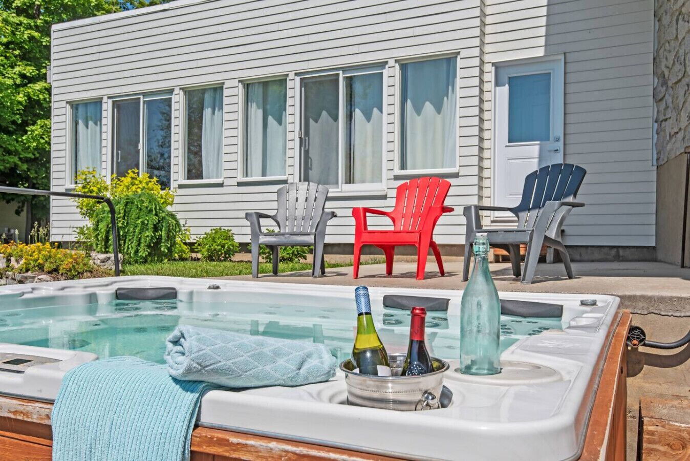 A hot tub with bottles and a towel, surrounded by outdoor chairs and a house in the background.