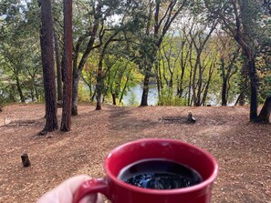 A hand holding a red mug with a dark liquid, possibly coffee or tea, in a forested area with a lake in the background. - Peaceful Rogue Riverfront Home on 3 Acres! (Grants Pass)