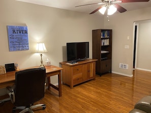 A room with a wooden desk, a chair, a television, a bookshelf, and a ceiling fan.