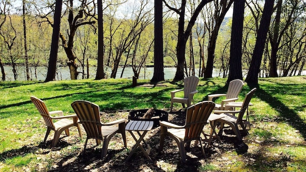 An outdoor seating area with Adirondack chairs around a fire pit, overlooking a lake with trees in the background.