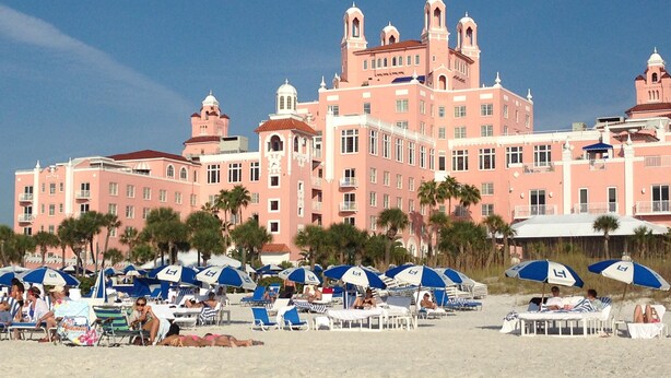 A large pink hotel with multiple towers and balconies, located on a beach with numerous beach umbrellas and lounge chairs.