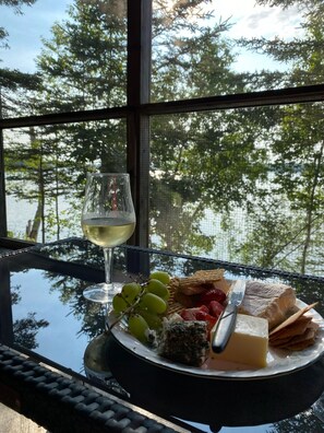 A glass of white wine, a plate with cheese, grapes, and crackers, and a basket of strawberries on a table by a window with a view of trees. - Driftwood Cottage; cozy oceanfront, 2 Kayaks, Wood Stove, Porch, Sunsets. (Brooksville)