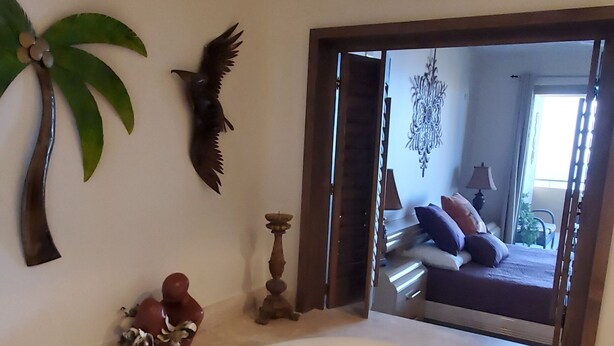Un bany amb una banyera blanca gran, una paret decorativa amb una palmera i un ocell, i vista cap a l'habitació per una porta oberta.