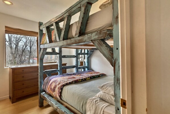 A bunk bed with a wooden frame, a window with a view of bare trees, and a wooden dresser.