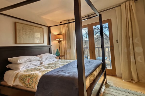 A four-poster bed with a patterned bedspread, a framed artwork above it, and a view of the outdoors through a glass door.