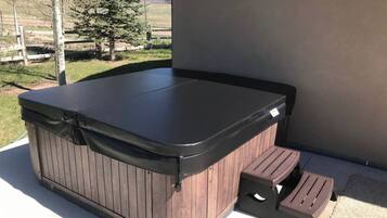 Un bain à remous avec une structure extérieure en bois et un bord supérieur noir, installé sur une terrasse offrant une vue sur une zone herbeuse et des arbres.