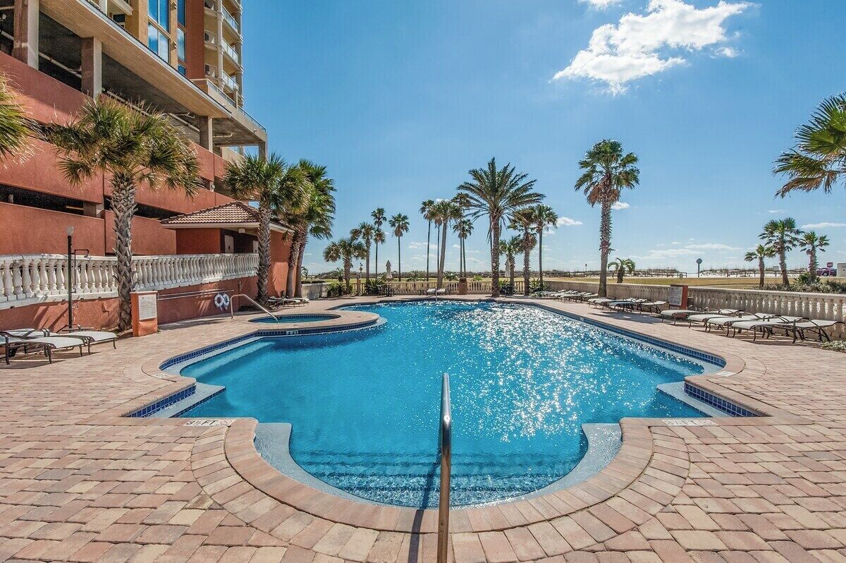 A swimming pool with a curved design, surrounded by palm trees and lounge chairs, with a building in the background.