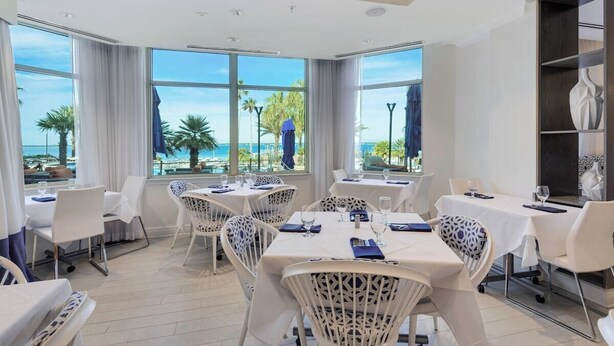 Un espace repas avec des tables et des chaises, de grandes fenêtres offrant une vue sur les palmiers et la mer, et un aménagement intérieur moderne.