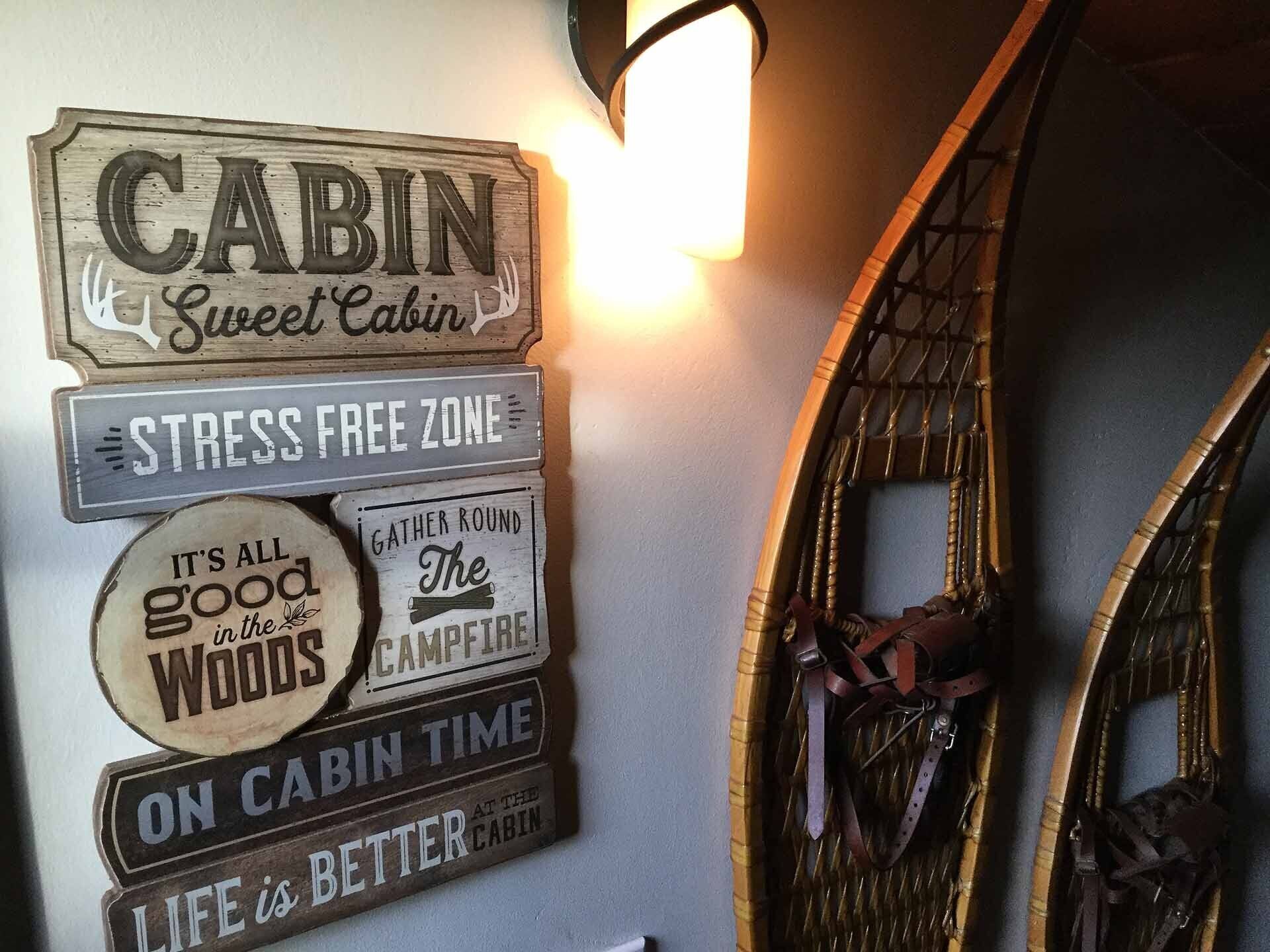 A wall with various signs including 'Cabin Sweet Cabin', 'Stress Free Zone', 'It's All Good in the Woods', 'Gather Round The Campfire', and 'Life is Better at the Cabin'.