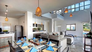 A spacious living area with a large sofa, dining table set for six, and a kitchen area with a refrigerator and cabinets. - Oceanfront Luxury Home Wifi 4 Smart TV'S Pool & Spa  (Puerto Morelos)