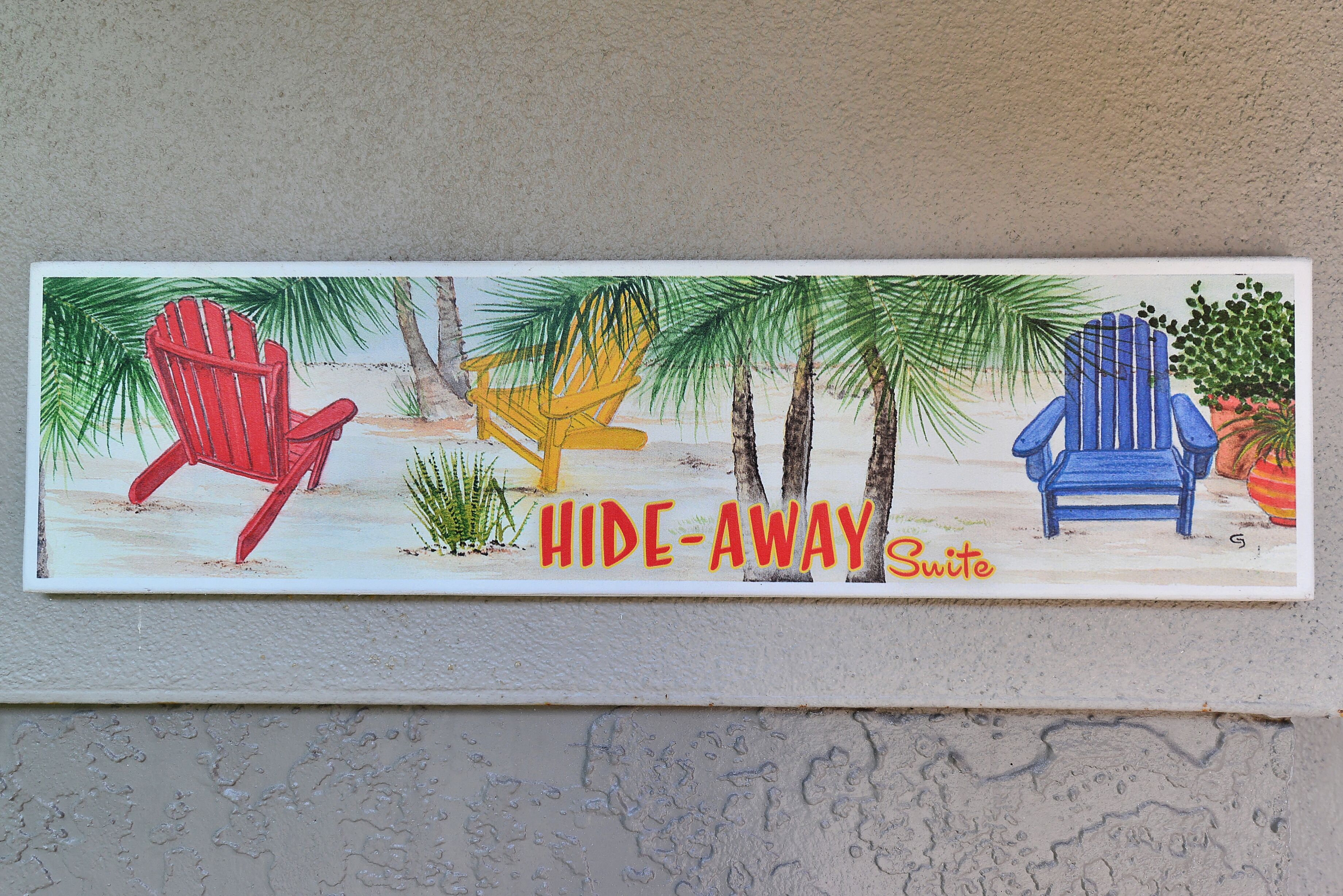 Et vægmonteret skilt med en illustration af et strandtema, der viser tre Adirondack-stole og palmer, med teksten "HIDE-AWAY Suite".