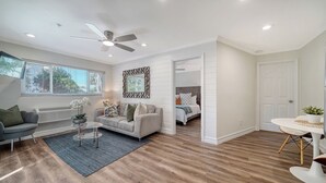 A spacious living room with a grey sofa, a coffee table, and a blue rug. There is a ceiling fan, a window with blinds, and a door leading to another room. -  🌷 Stunning 1-Bedroom Apartment Overlooking Pool, steps to Siesta Beach (Siesta Key)