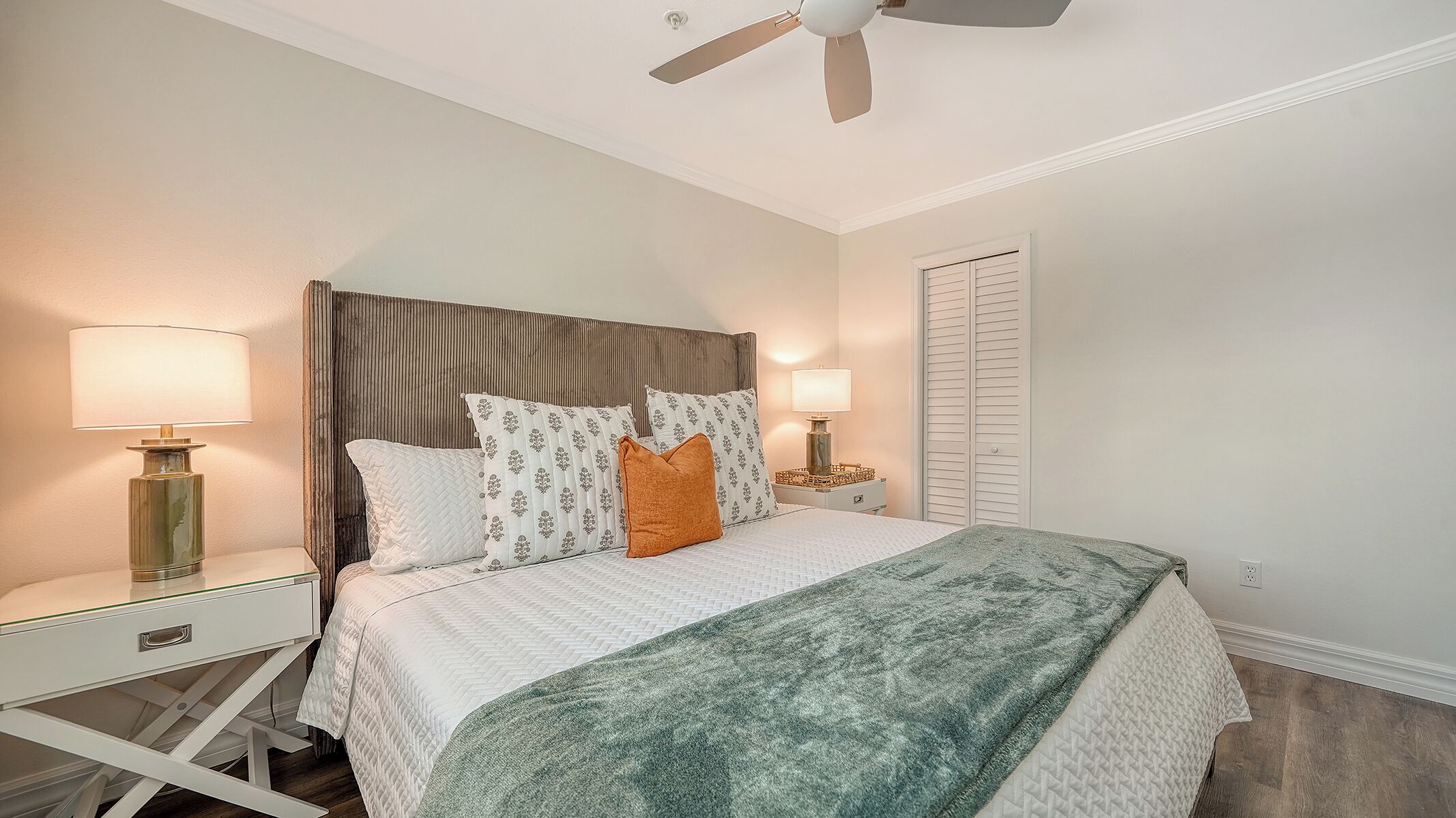 A neatly arranged bedroom with a bed, two bedside tables with lamps, a ceiling fan, and a closet with sliding doors.