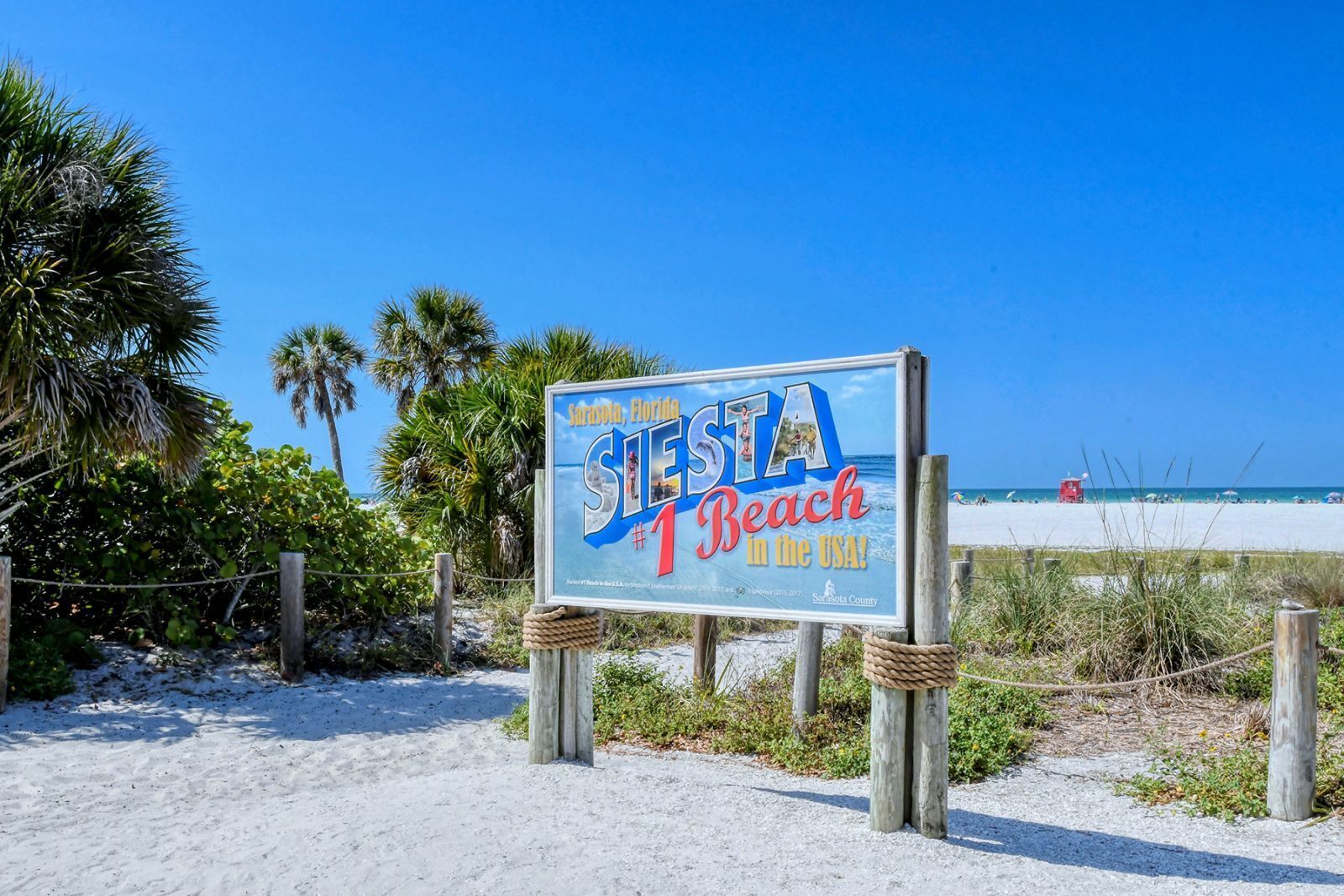 Ein Strandschild mit der Aufschrift 'Sarasota, Florida – der beste Strand der USA!', mit freiem Blick auf den Strand und das Meer im Hintergrund.
