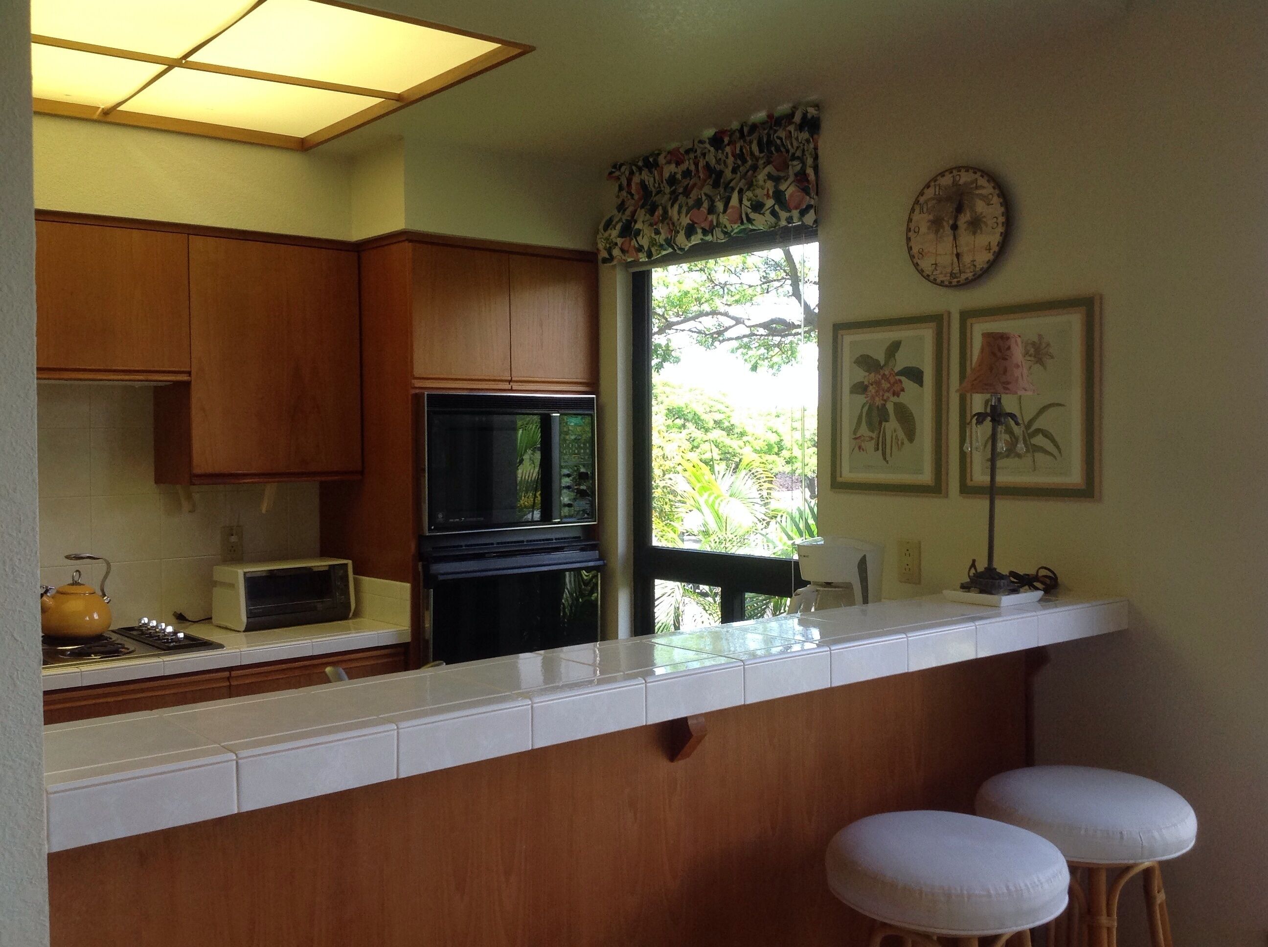 A kitchen with wooden cabinets, a microwave, a stove, and a window with a view of greenery. There are two white stools and a clock on the wall.