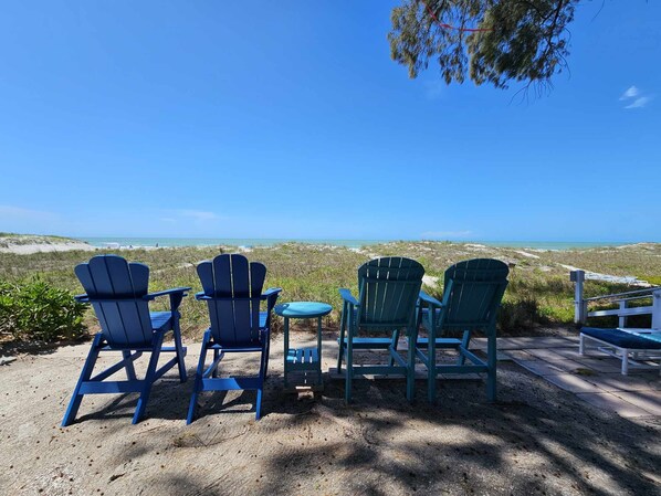 A beachside seating area with Adirondack chairs and a small table, overlooking a sandy beach and ocean.