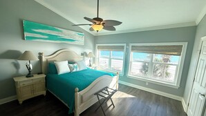 Quarto com cama, mesas de cabeceira, ventilador de teto e vista para a praia através de amplas janelas.
