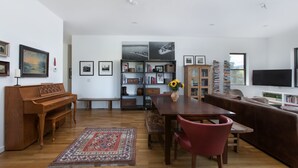 Een ruime woonkamer met een houten piano, een eettafel met stoelen, een rode bank, een boekenkast en een grote televisie.
