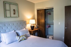 A bedroom with a tufted headboard, a bedside table with a lamp, and a wardrobe with hanging clothes.