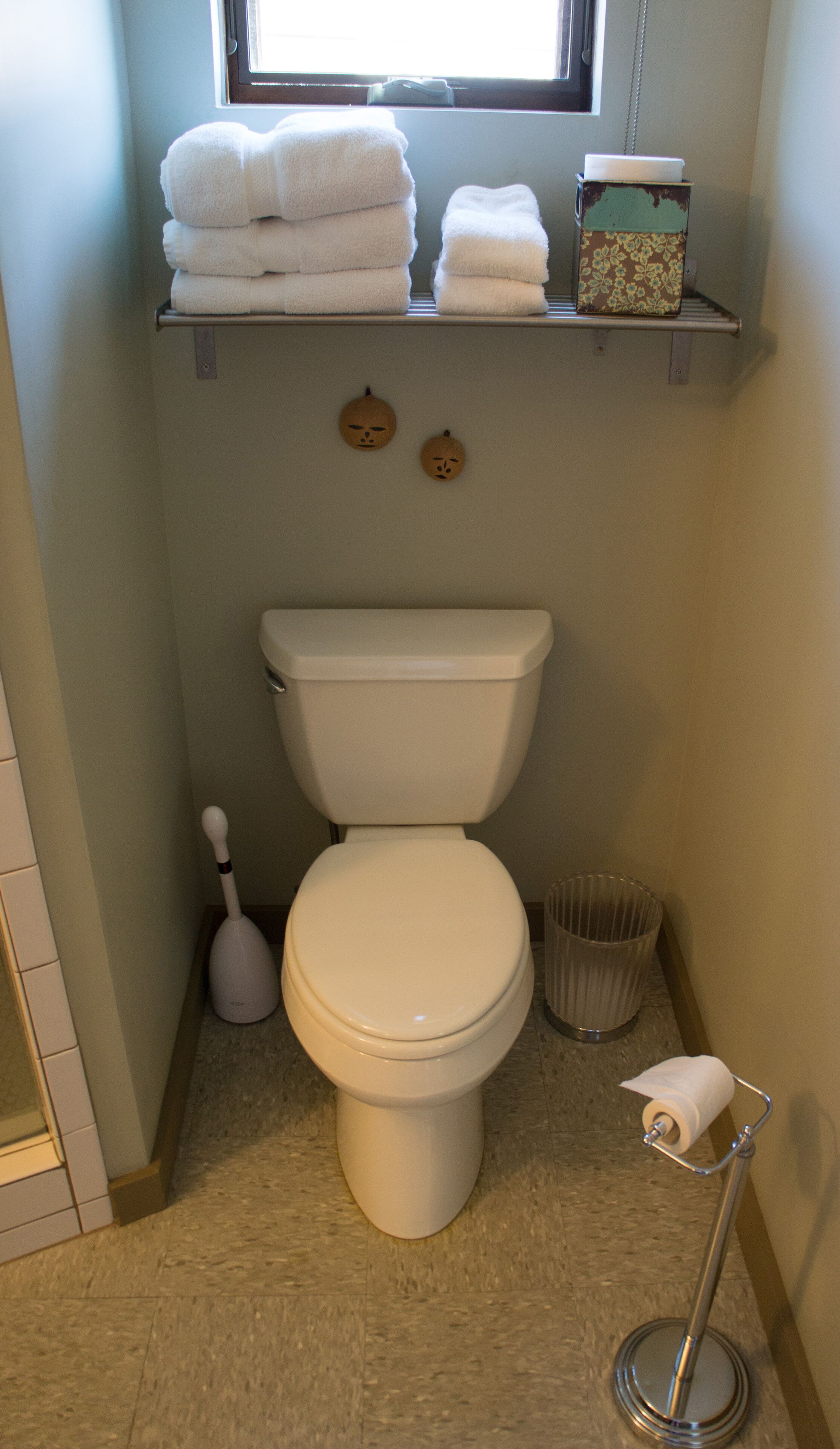 Un baño con inodoro, escobilla, cubo de basura y estante con toallas.