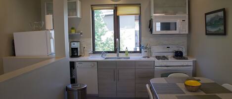 A compact kitchen with a microwave, stove, and sink. There is a clock on the wall and a window with blinds.